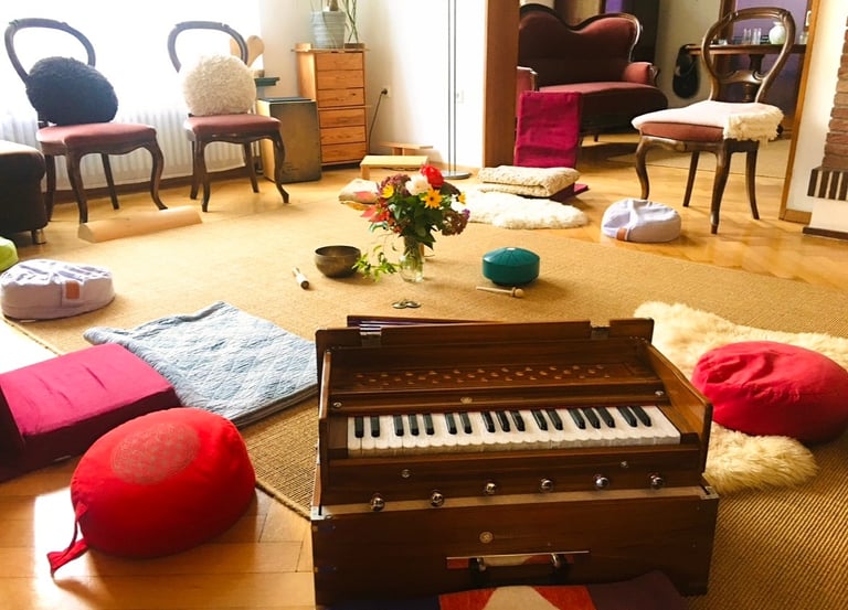 ein Raum mit Stuhlkreis und Sitzkissen, einem Harmonium und Blumenstrauß in der Mitte