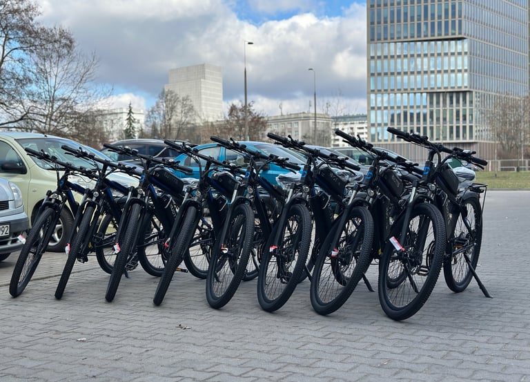 A row of black electric mountain bikes parked on a city pavement with modern office buildings in the background.