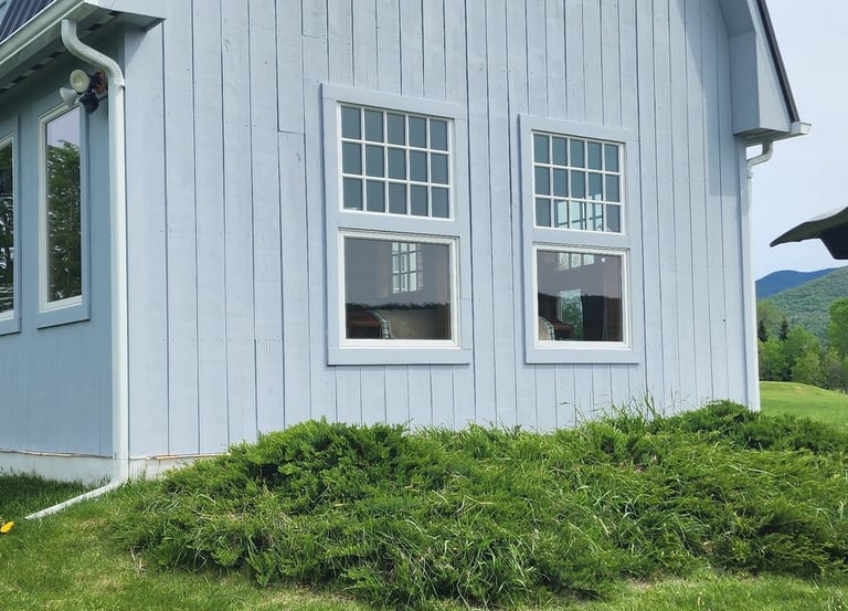A light blue gambrel roof barn conversion home with white windows and a green lawn with dandelions.