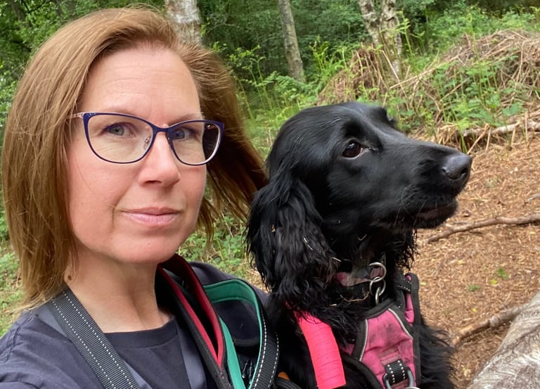 A dog trainer and her black cocker spaniel sit together on a log during a walk