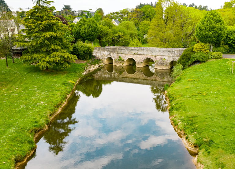 Vue aérienne d'un vieux pont en arc de pierre enjambant une rivière calme, entouré d'une végétation 