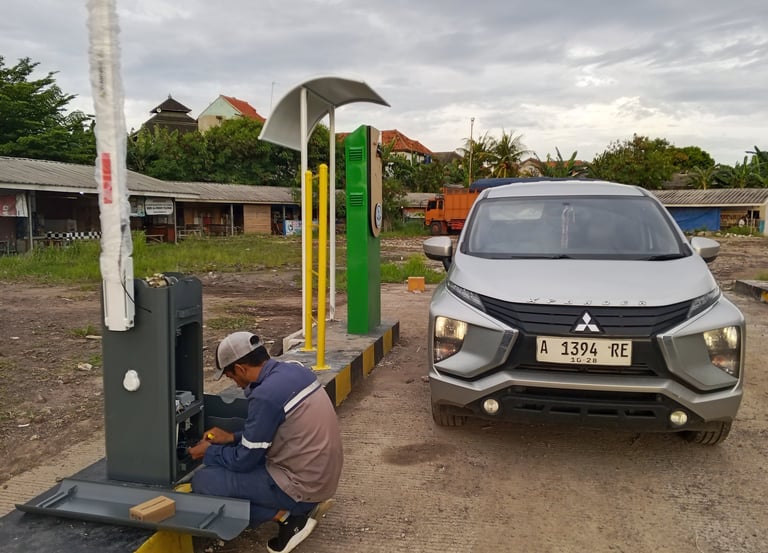 A technician repairs a parking lot boom gate system next to a silver Mitsubishi Xpander car.