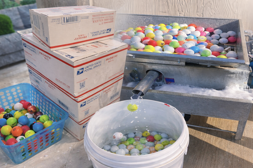 Used golf balls being cleaned in a sorting machine next to USPS Priority Mail shipping boxes.