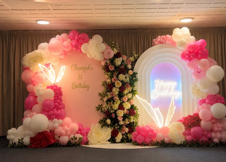 Elegant first birthday backdrop with pink balloon arch, white floral accents, and neon birthday signs.