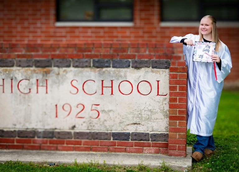 A blonde high school graduate in a white cap and gown posing by a brick school sign.