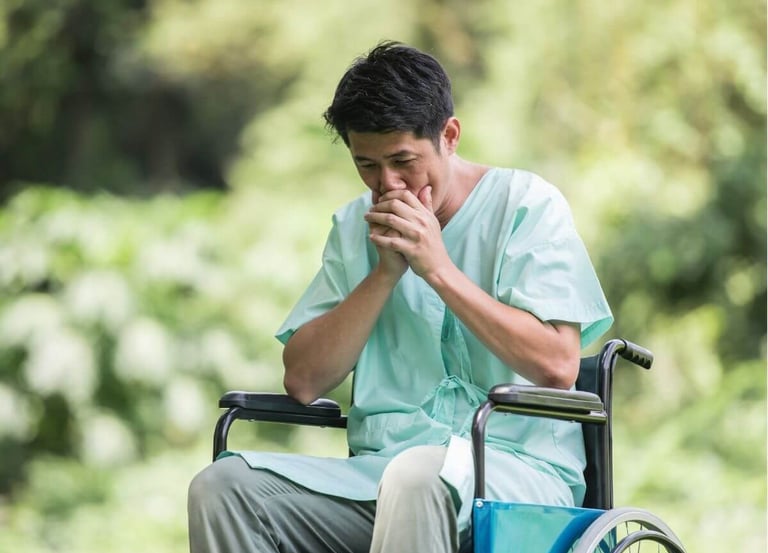 A patient in a hospital gown sitting in a wheelchair outdoors, looking pensive and concerned.
