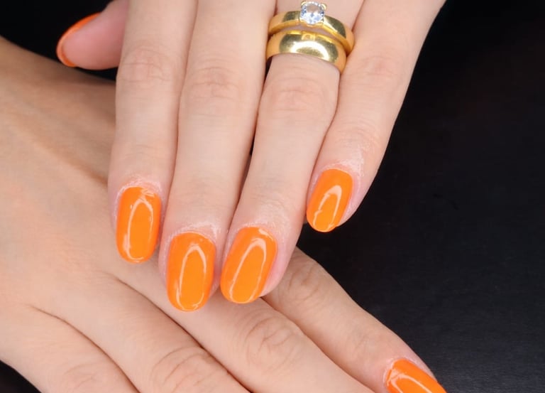 Professional manicure featuring vibrant orange gel nail polish and gold rings on a woman's hands.