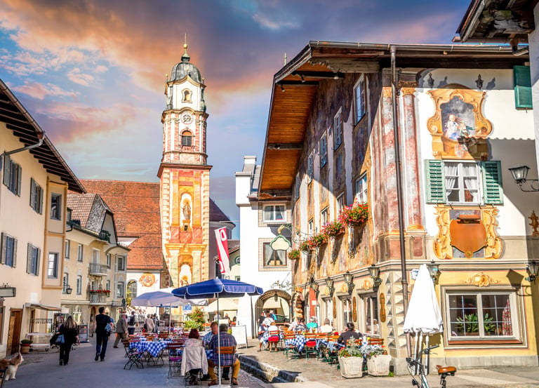 Charming Mittenwald street scene with Lüftlmalerei murals, outdoor cafes, and the St. Peter and Paul church tower.