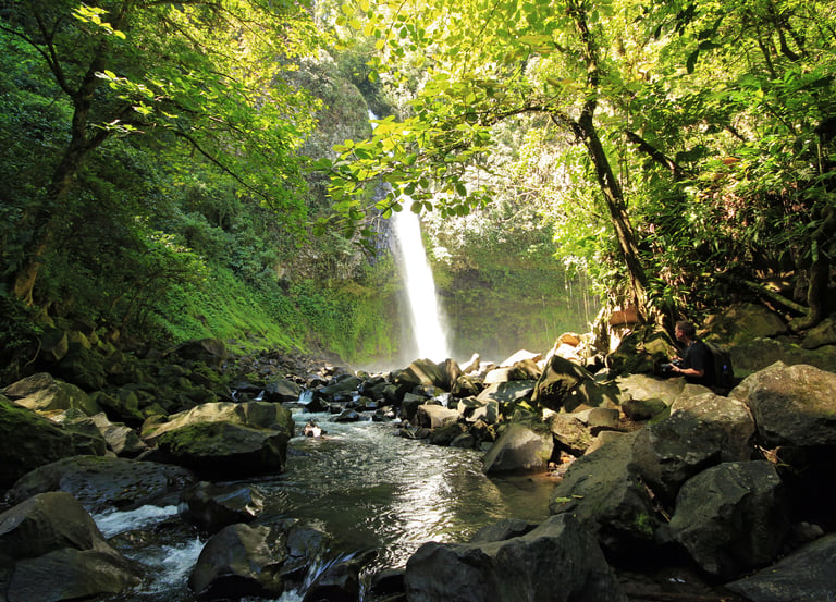 A scenic waterfall flowing into a rocky riverbed surrounded by a lush green tropical forest.