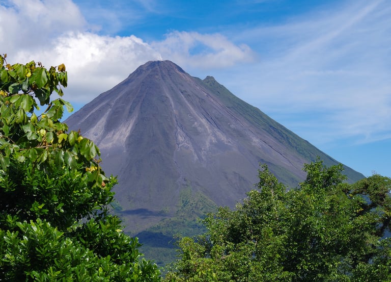 Arenal Volcano in Costa Rica with lush green tropical trees and a blue sky with white clouds.
