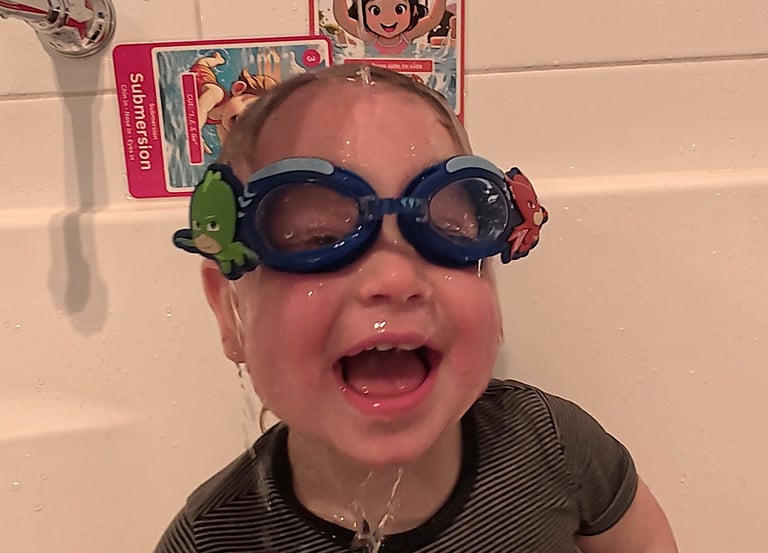 Baby sitting in the bath with goggles with water over the face and swim skill flash cards stuck on the wall.