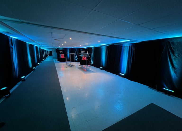 Professional event space with black drapes, blue LED uplighting, and clothing racks in a wide hallway.