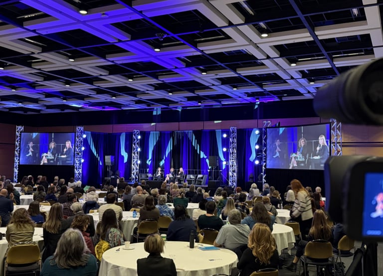Audience at a professional conference seated at round tables watching a panel discussion on large screens.