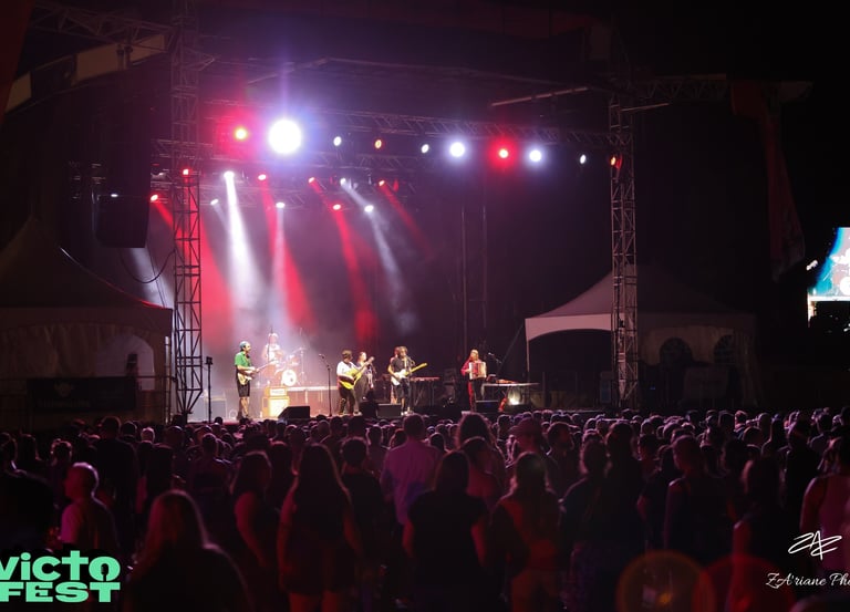 A live band performs on a brightly lit outdoor stage for a large crowd at VictoFest music festival.