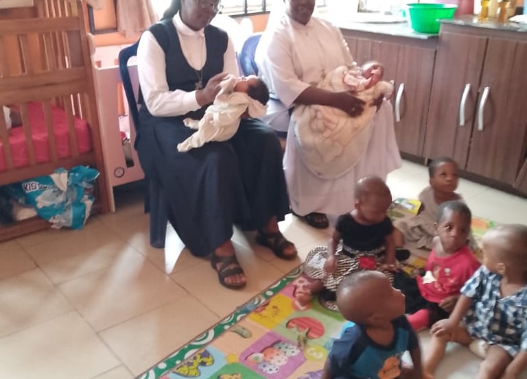 Two Catholic nuns holding infants while sitting near toddlers on a play mat in a childcare nursery.