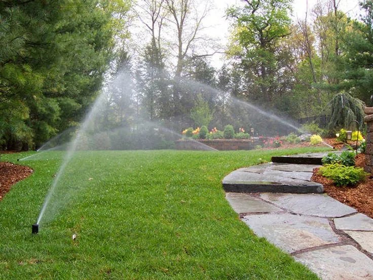 Automatic underground sprinkler system watering a large residential backyard with stone pathways.