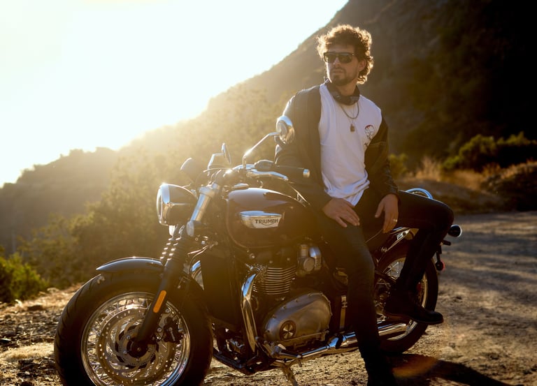 Hombre con gafas de sol y chaqueta de cuero, sentado en una motocicleta Triumph, al atardecer en un paisaje montañoso