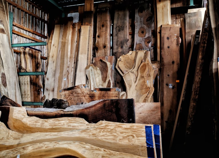 Assorted live edge wood slabs and reclaimed timber boards stacked in a woodworking lumber yard warehouse.