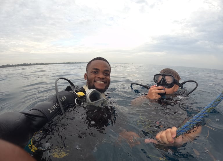 A smiling man and young boy wearing scuba diving gear floating on the ocean surface during a dive excursion.