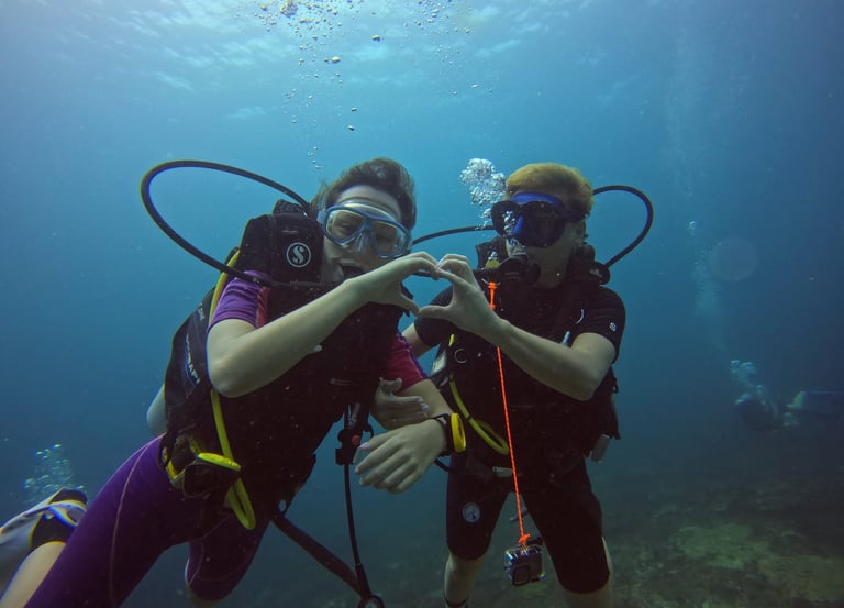 Two scuba divers making a heart shape with their hands while underwater diving in clear blue ocean water.