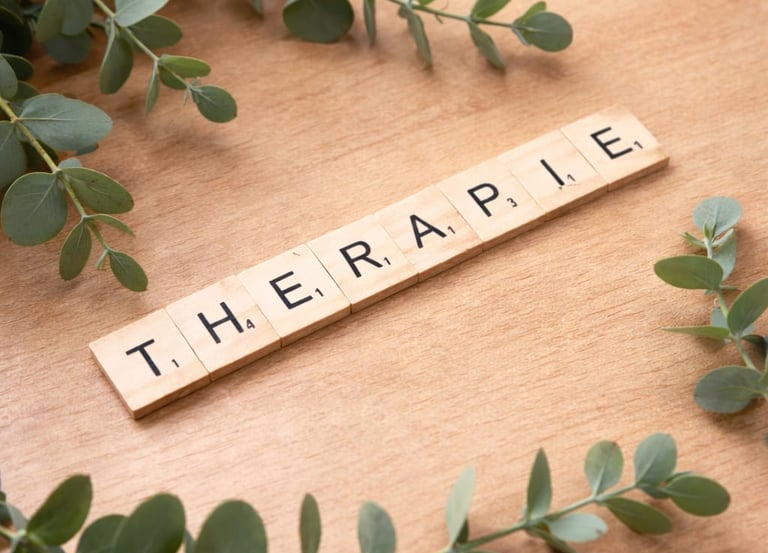 Wooden letter tiles spelling Therapie surrounded by green eucalyptus leaves on a desk.