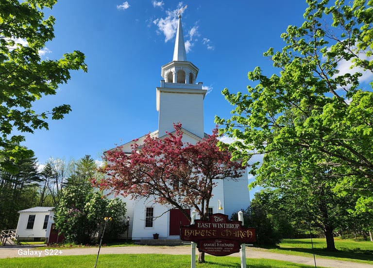 Historic East Winthrop Baptist Church in Maine with a white steeple and blooming pink crabapple tree.