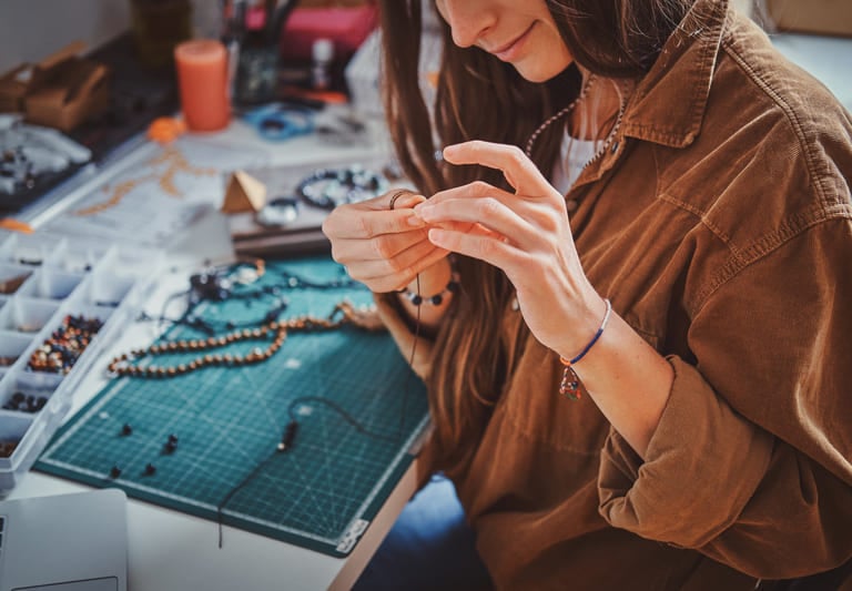 Artesana creando collares hechos a mano con cuentas y herramientas de joyería sobre mesa de trabajo.