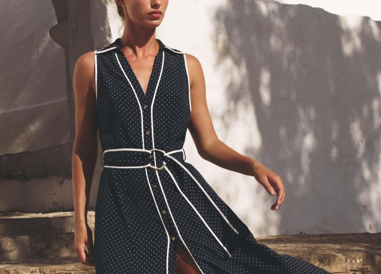 A woman in a navy blue polka dot midi dress with white trim posing outdoors near a stone wall.