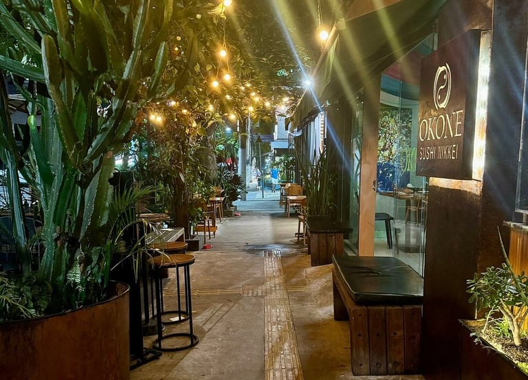Entrada do Okone Sushi Nikkei à noite, com fachada em madeira, placa iluminada e luzes decorativas em rua arborizada.