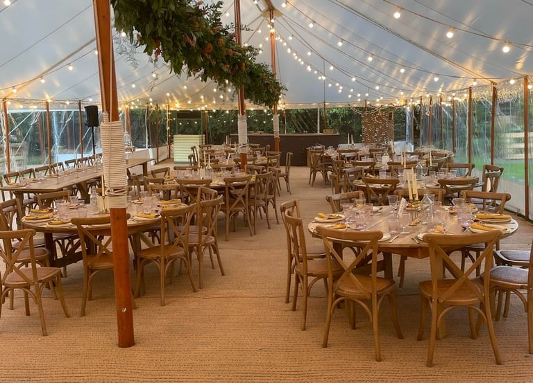 Elegant outdoor wedding reception tent with round wooden tables, bistro lighting, and floral greenery.