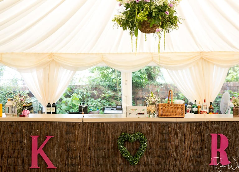 Rustic wedding bar setup with pink initials and a heart wreath under a white marquee tent.