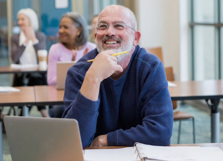 Mature man learner in a class wearing glasses smiling with laptop and mature women background