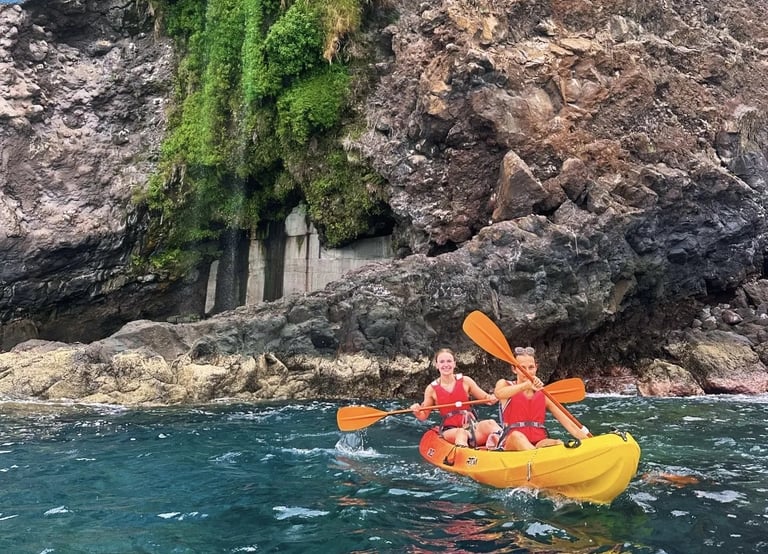 Two women in lifejackets paddling a yellow kayak past a green cliff waterfall on the Madeira coast