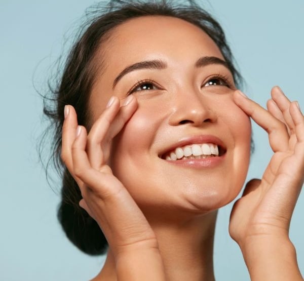 Mujer sonriente con piel radiante aplicando crema para los ojos en su rutina de hidratación facial.