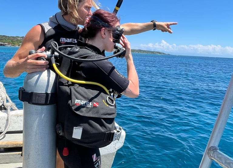 A scuba diving instructor points to the ocean while helping a student diver prepare for a boat dive.