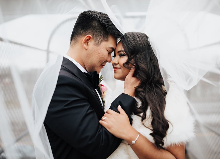 A smiling bride and groom embracing under a flowing white wedding veil during a romantic outdoor ceremony.