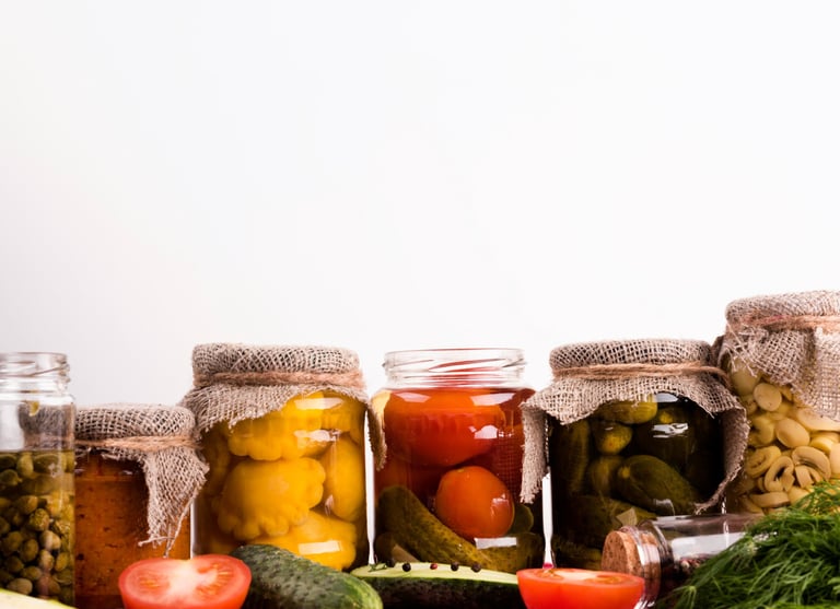 Assorted glass jars of pickled vegetables including cucumbers, tomatoes, and squash on a red checkered cloth.