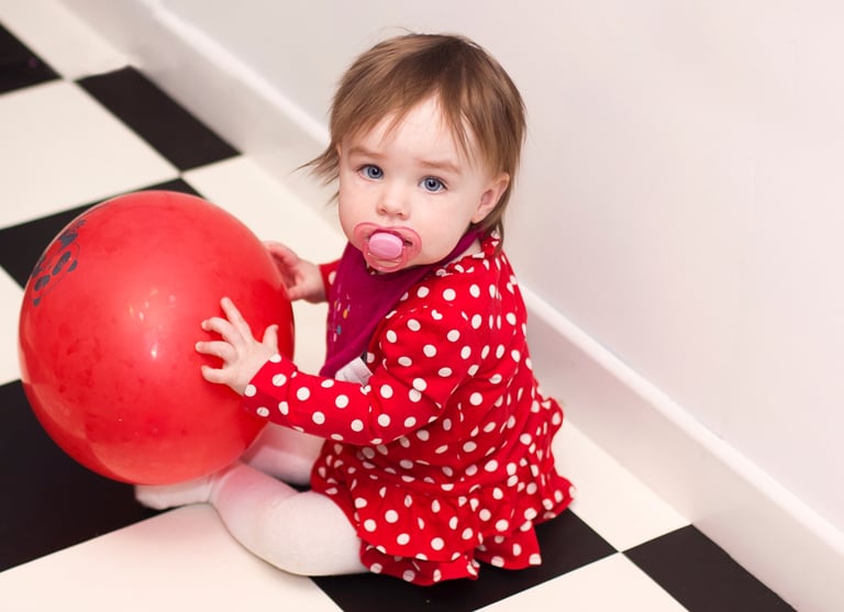 A toddler in a red polka dot dress holding a large red ball on a black and white tiled floor.