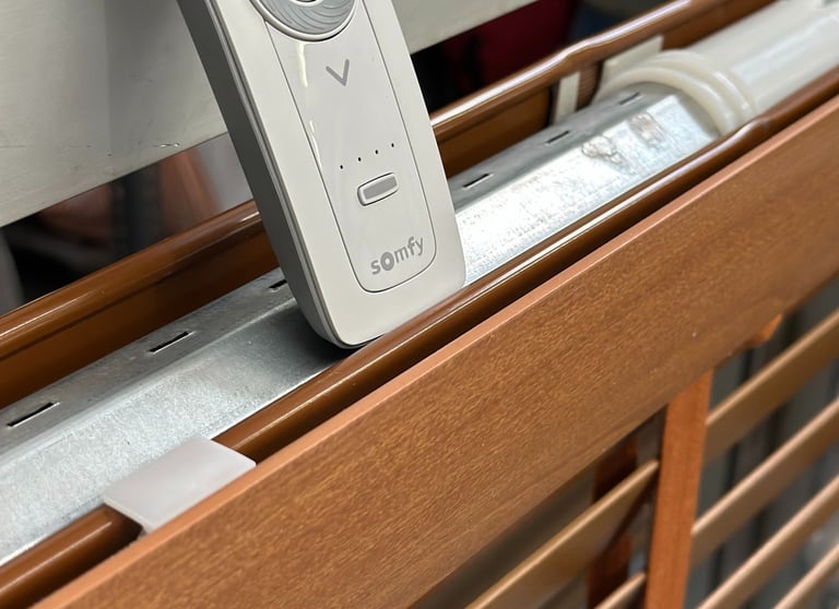 Somfy remote control resting on motorized wooden window blinds with metal hardware.