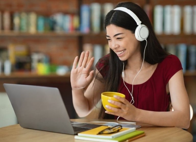 Female student learning French online, wearing headset, smiling and waving at her laptop