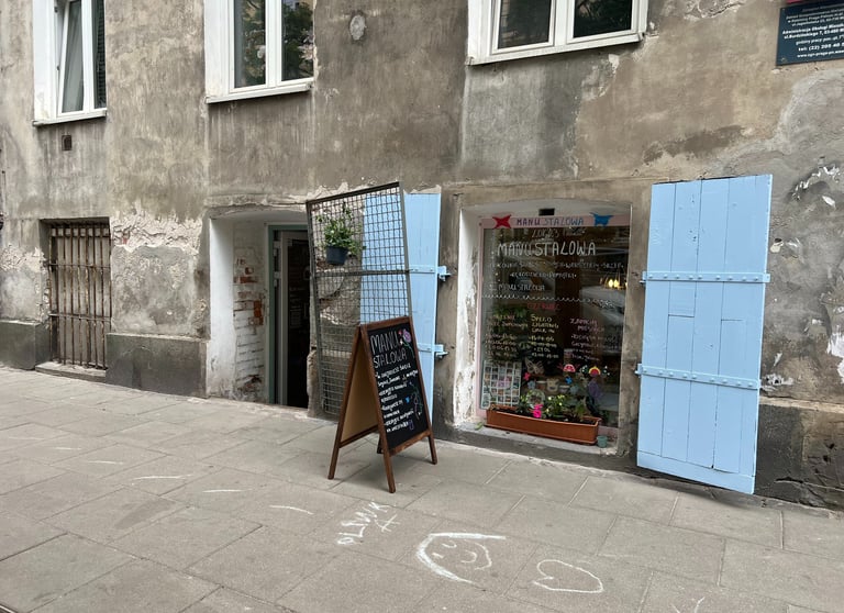 Manu Stalowa storefront in Warsaw with blue shutters and a chalkboard menu on a cobblestone sidewalk.