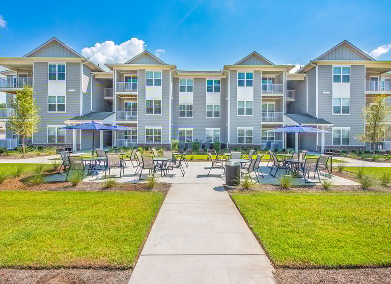 Ground-level view of outdoor patio seating with umbrellas between apartment buildings at Arrogate Village in Summerville, SC