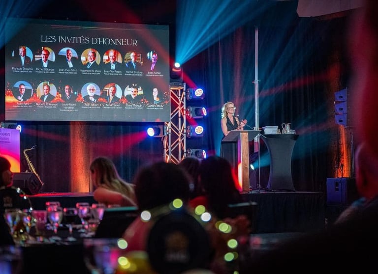 A female speaker at a gala event podium with an honorary guest display screen and stage lighting.