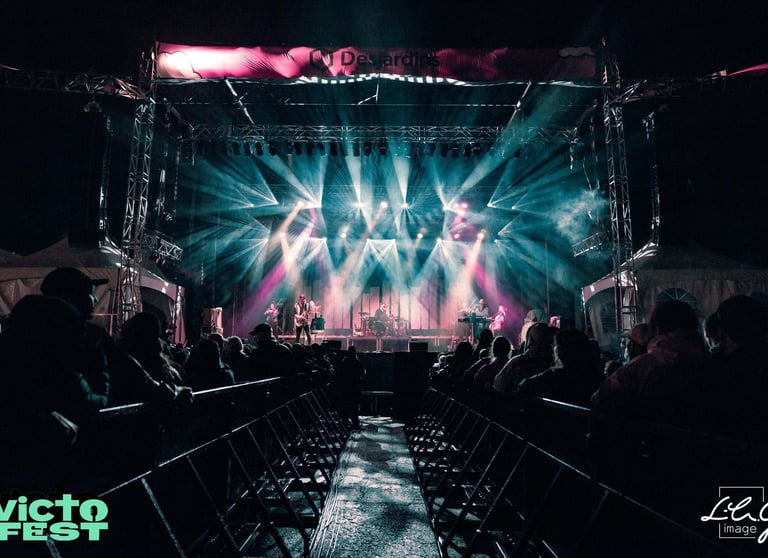 A band performs on a brightly lit outdoor stage with dramatic spotlights at VictoFest music festival.