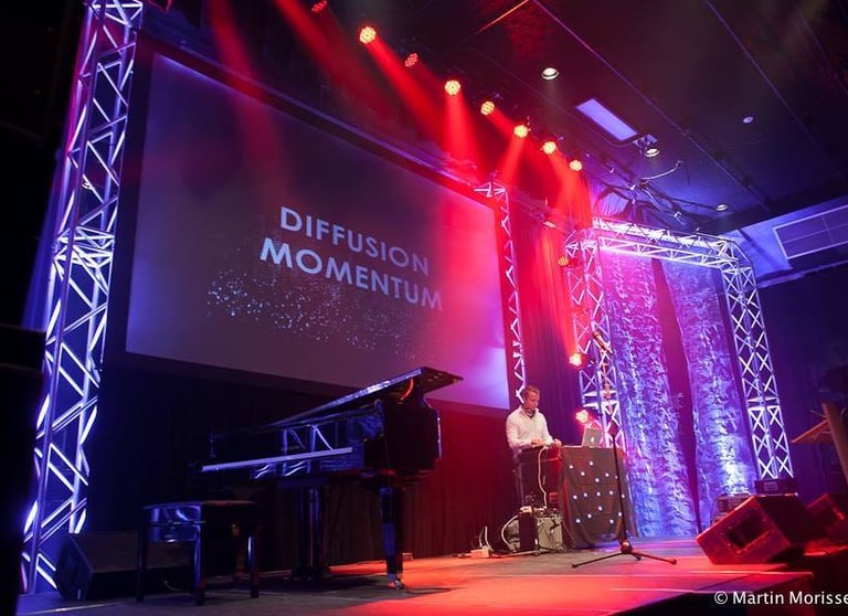 Live performance on stage featuring a grand piano and DJ with red and blue concert lighting.