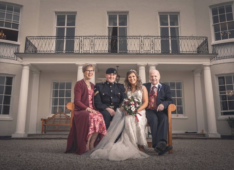 A bride and groom in military uniform sit with family outside a white manor wedding venue.