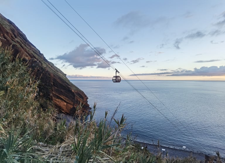 A scenic cable car descending a coastal cliff over the blue Atlantic Ocean in Madeira at sunset.
