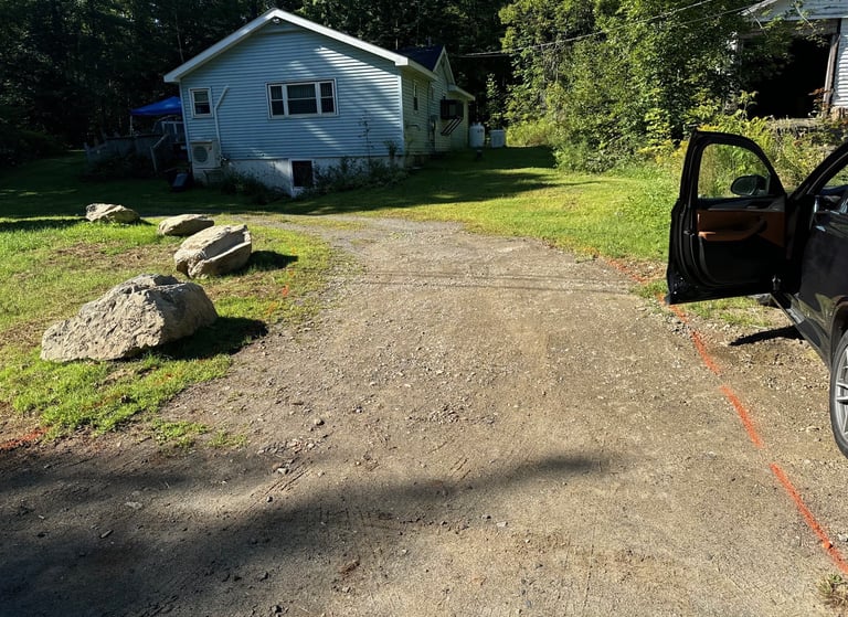 A gravel driveway leading to a light blue house surrounded by green trees and large decorative landscape rocks.