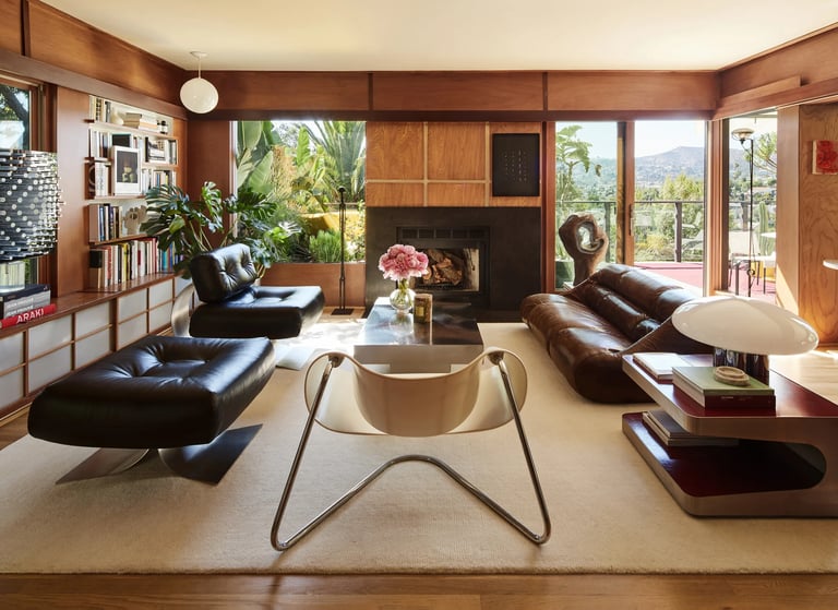 Modern living room with leather sofas, fireplace, and large windows overlooking greenery.