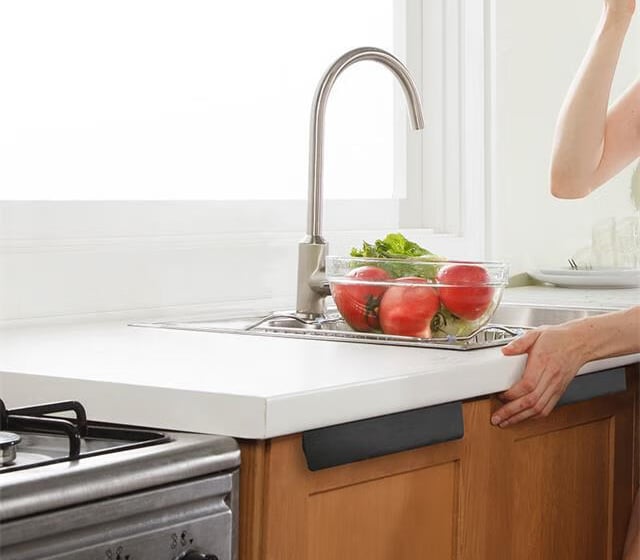 Stainless steel faucet over kitchen sink with fresh tomatoes and lettuce, person adjusting light countertop nearby